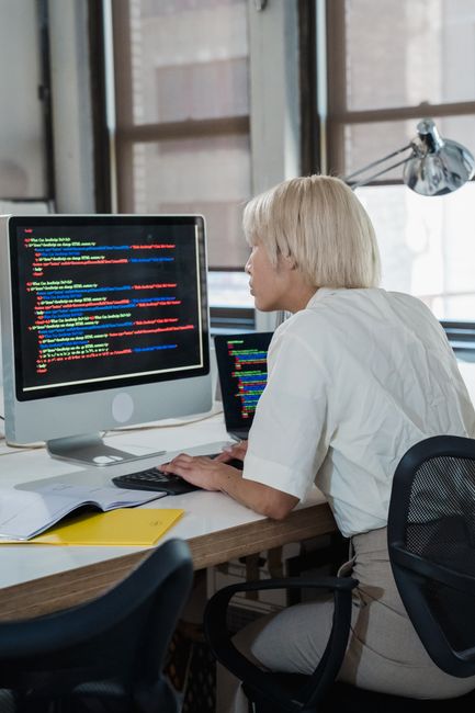 A person with short blonde hair and wearing a white shirt is seated at a desk in a well-lit room with large windows, working on a computer with a large monitor displaying lines of colored code. The de