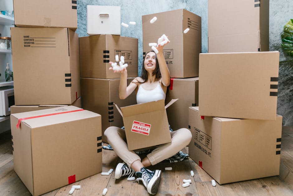 A man with a joyful expression is inside a bright living room, carrying a large white textured vase with green leafy plants in one hand and a small cardboard box labeled 'FRAGILE' in the other. The ro