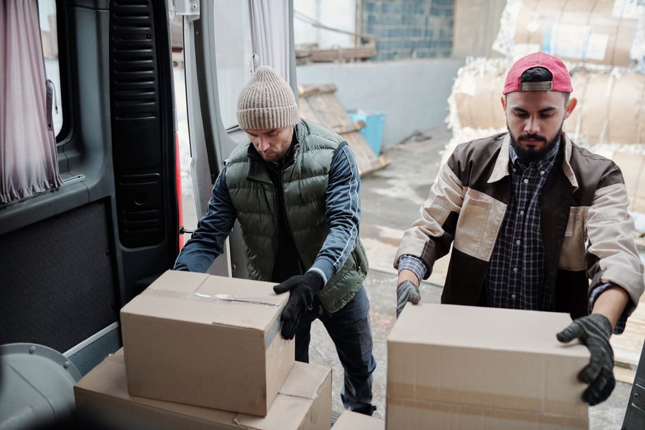 The image shows the side view of a white commercial van with its rear doors open, revealing a spacious interior packed with numerous moving boxes and packaging materials. Several cardboard boxes of va